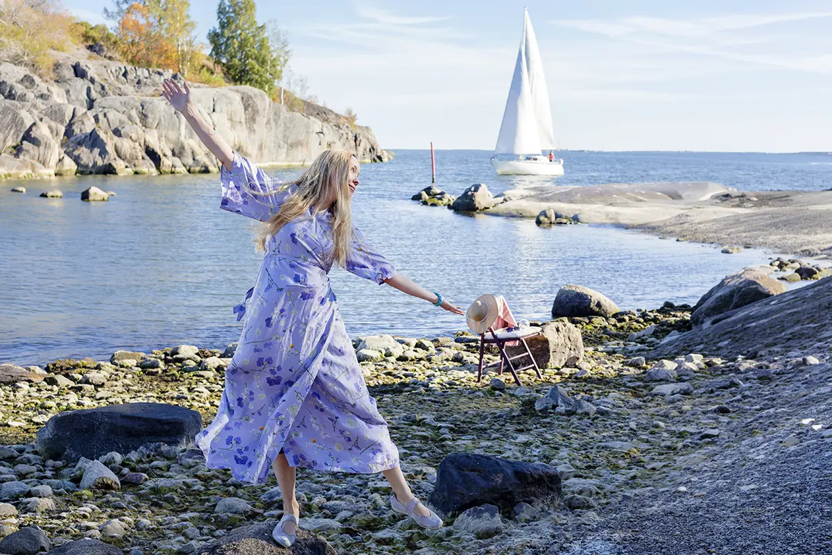 Studio Aina. Nainen lilassa mekossa tasapainoilee leikkisästi kivikkoisella rannalla merimaisemassa. Taustalla purjevene ja kallioita.